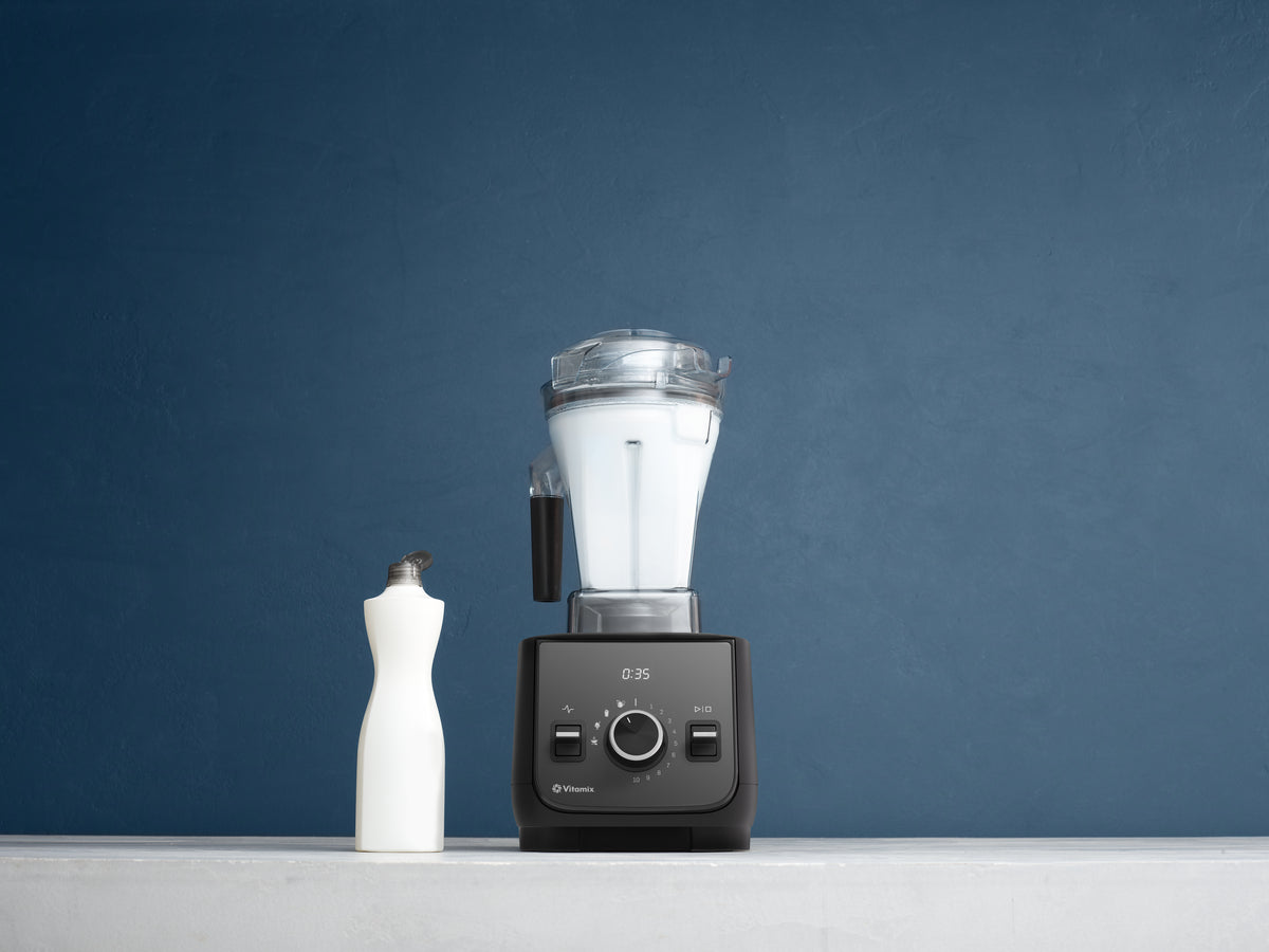 A Vitamix Ascent X2 professional blender with a clear container with white bubbles inside and black base, shown next to a white bottle of detergent on a light grey surface against a dark blue textured background.