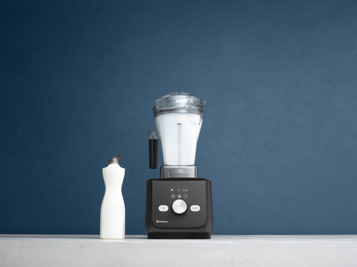 A Vitamix Ascent X blender with a black base and clear container, filled with white bubble liquid, standing on a light gray surface against a dark blue textured background. A white bottle of washing up liquid is positioned to the left of the blender.