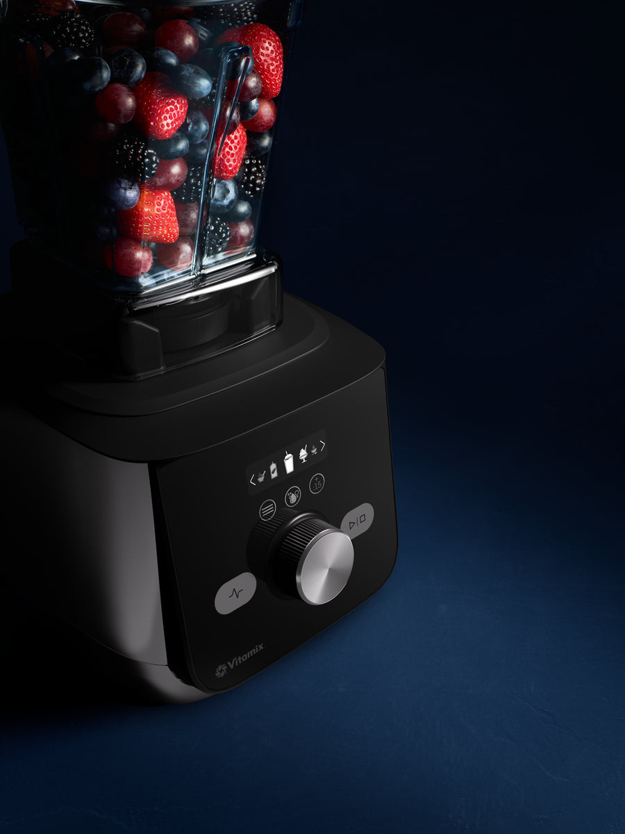 A Vitamix blender with a black base and clear container jug filled with mixed berries, strawberries, and blueberries, photographed on a dark blue background.