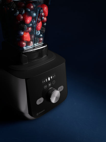 A Vitamix blender with a black base and clear container jug filled with mixed berries, strawberries, and blueberries, photographed on a dark blue background.