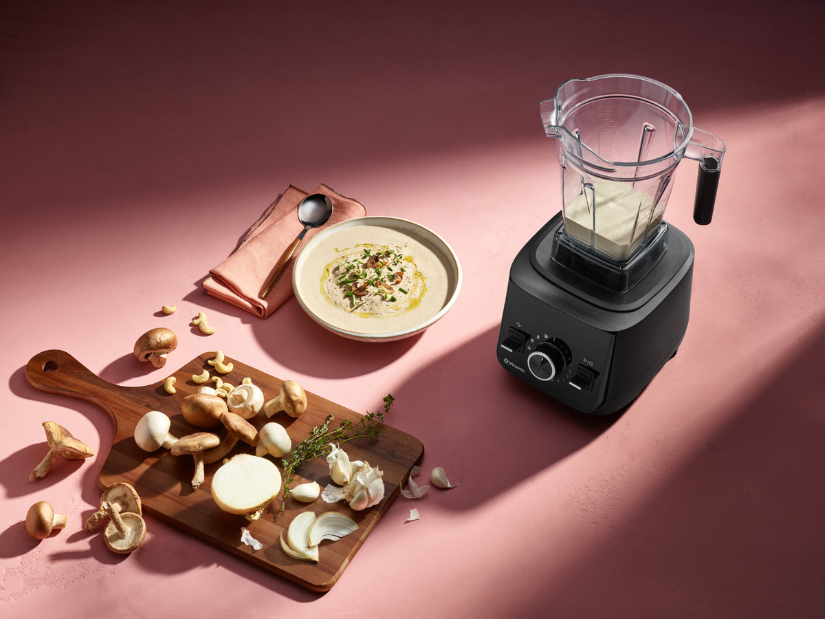 Ascent X2 blender filled with a creamy beige liquid stands on a pink surface beside a bowl of soup, mushrooms on a wooden cutting board, garlic cloves, and a folded pink cloth napkin with a spoon. The scene is styled in a real kitchen-like setup with strong directional light and shadows.