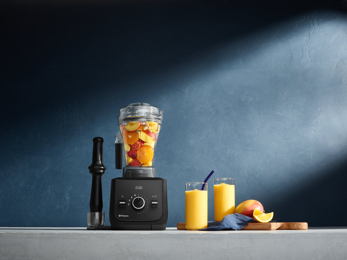 Ascent X2 blender full of yellow and red fruit on a light surface with blue background and a tamper beside it. To the right is two glasses of yellow smoothie, a chopping board, a mango slice of lemon and a blue napkin.