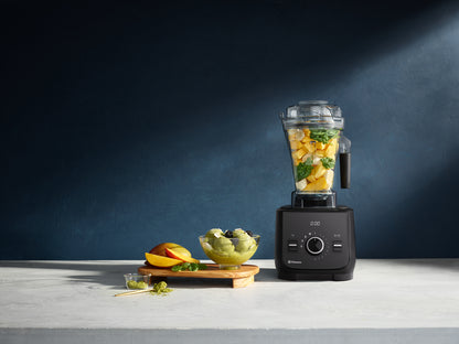 A black Vitamix Ascent X2 blender filled with chopped fruit and green leaves stands on a light stone countertop against a dark blue wall. A small bowl of green Macha ice cream, a sliced mango, and a few mint leaves sit beside it as styled props.