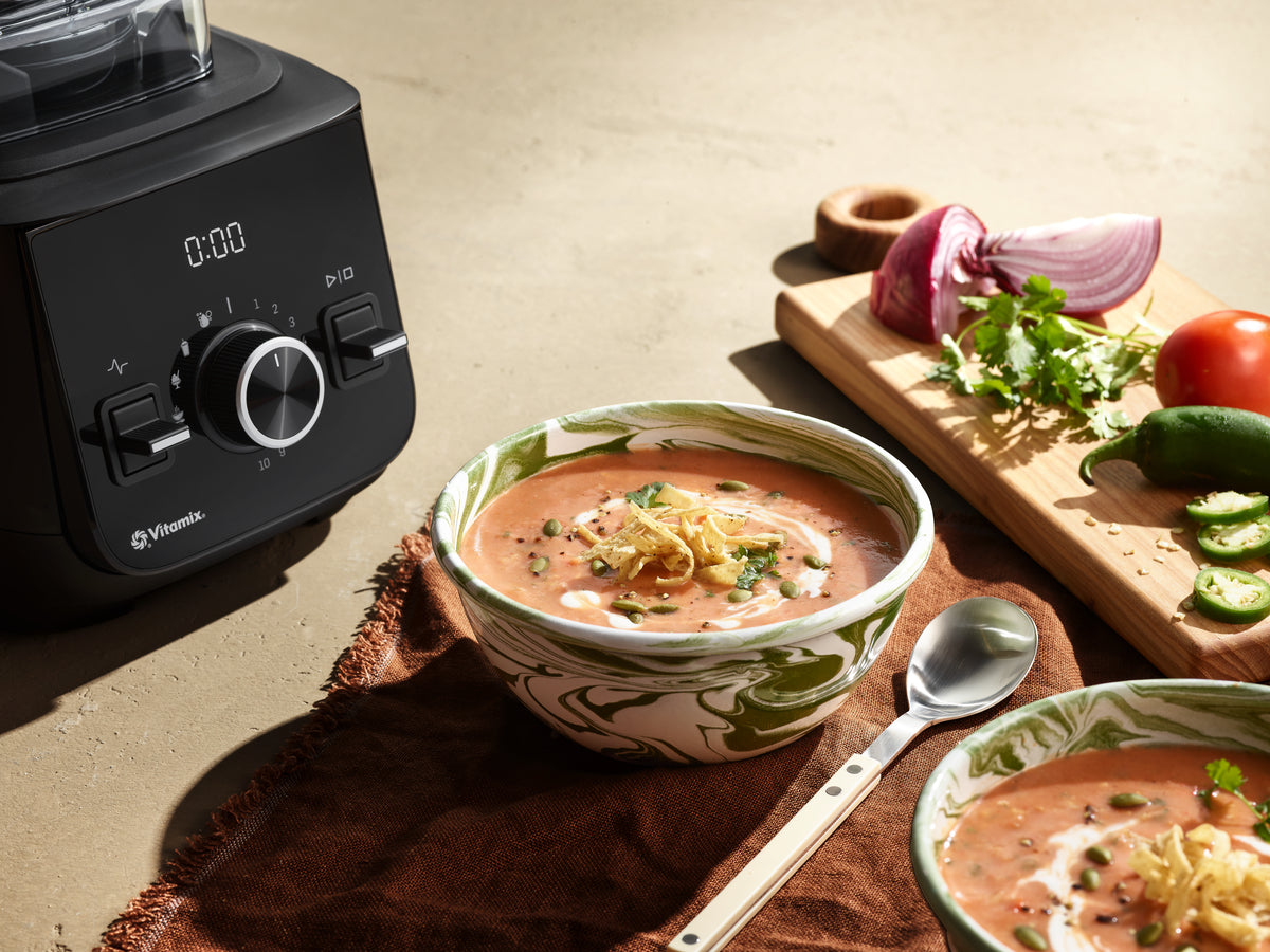 Vitamix Ascent X2 blender in black is shown beside bowls of prepared soup on a warm beige tabletop, with a spoon, cutting board, red onion, tomato, jalapeño, and corriander nearby.