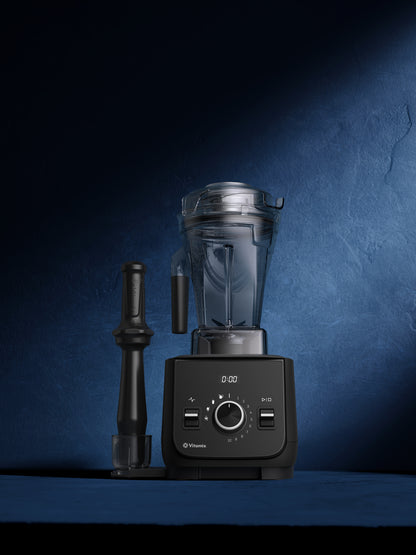 A Vitamix X2 blender with a clear container and black base, shown alongside a black tamper on a dark blue textured background.
