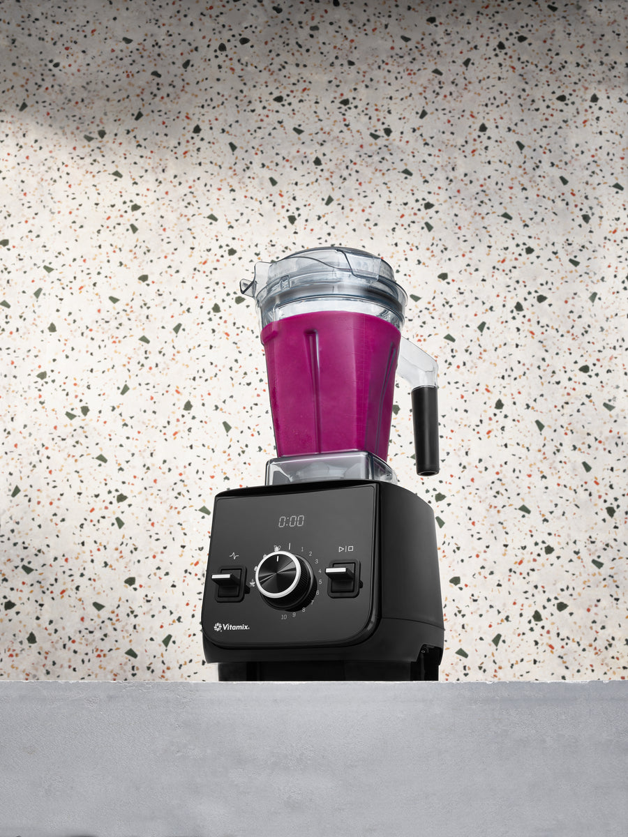 A Vitamix Ascent X2 blender with a black base and a clear container filled with magenta-purple liquid, set against a speckled terrazzo-style wall and a grey countertop.