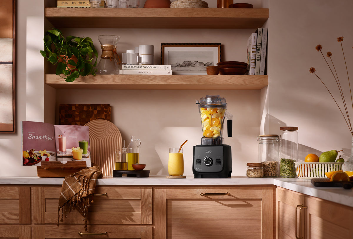 A black Ascent X2 Vitamix blender filled with chopped yellow fruit sits on a kitchen counter in a warm, styled kitchen setting with wooden shelves, glass jars, a smoothie recipe book and a yellow drink nearby.