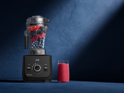 A Vitamix Ascent X blender with a clear container filled with mixed berries sits on a dark blue surface against a textured blue wall, with a glass of pink smoothie beside it.