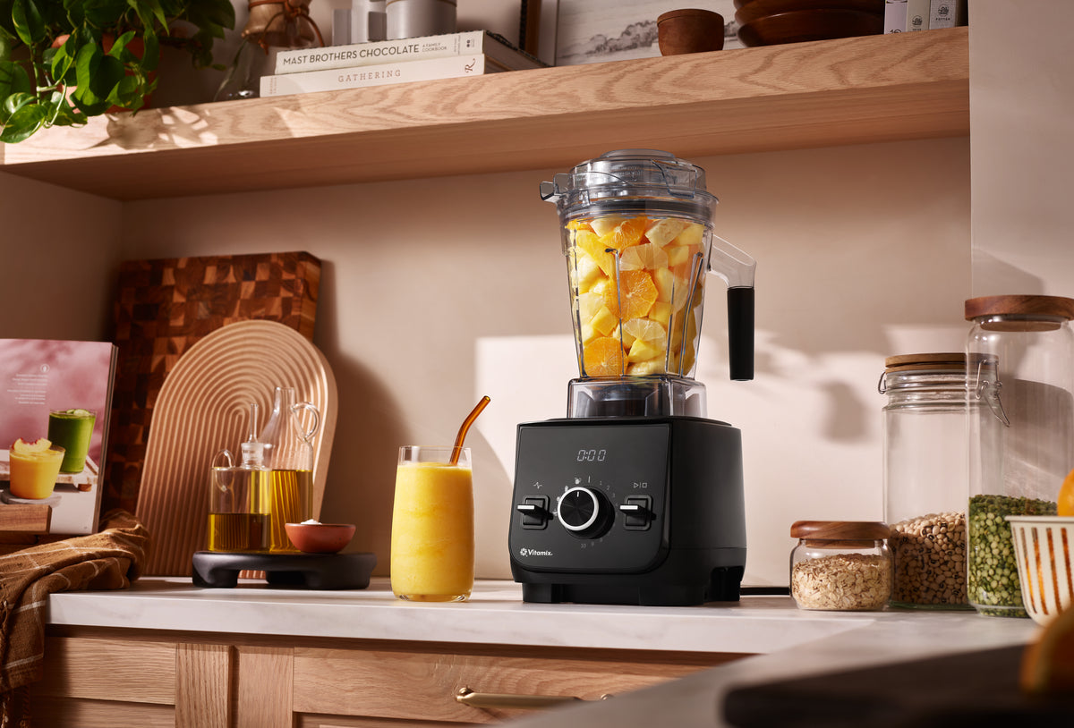 A black Ascent X2 Vitamix blender filled with chopped yellow fruit sits on a kitchen counter in a warm, styled kitchen setting with wooden shelves, glass jars, and a yellow drink nearby.