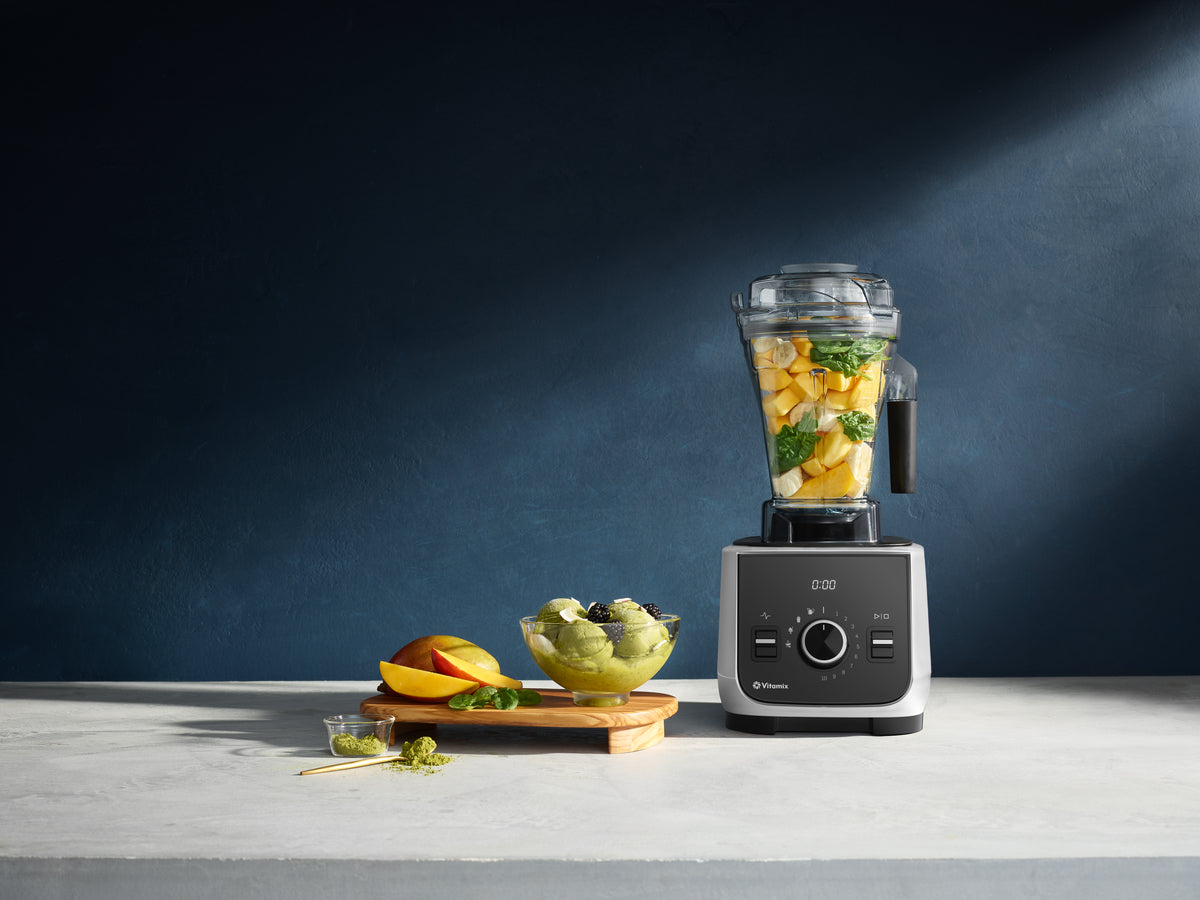 Ascent X2 countertop blender filled with chopped fruit and greens stands on a light stone surface against a dark blue wall. A small bowl of green matcha & fruit  icecream a sliced mango, and a few mint leaves sit beside it as styled props.