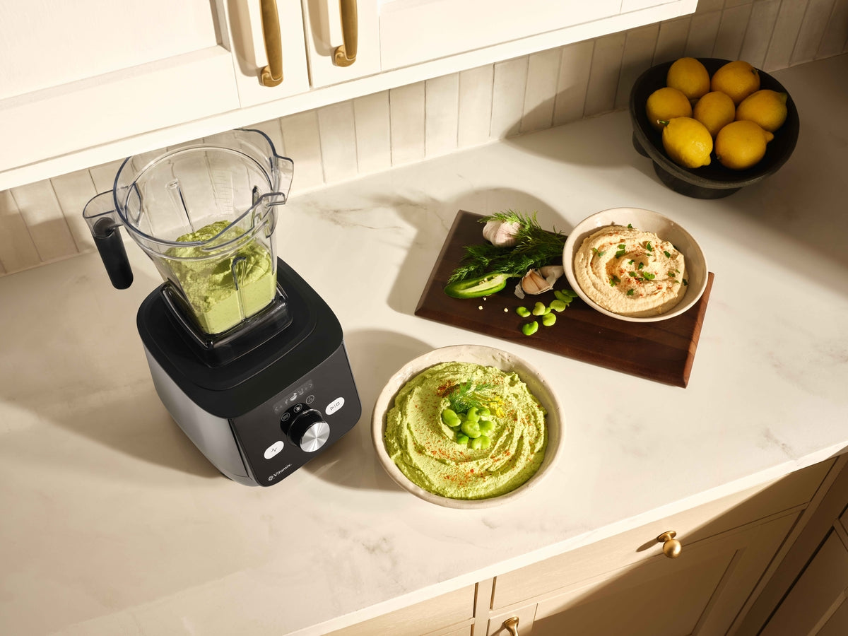 A countertop kitchen scene showing a black Ascent X blender with a clear container, alongside bowls of green dip and hummus, a wooden board with fava beans and garlic, and a bowl of lemons.