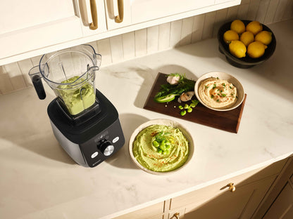 A countertop kitchen scene showing a black Ascent X blender with a clear container, alongside bowls of green dip and hummus, a wooden board with fava beans and garlic, and a bowl of lemons.