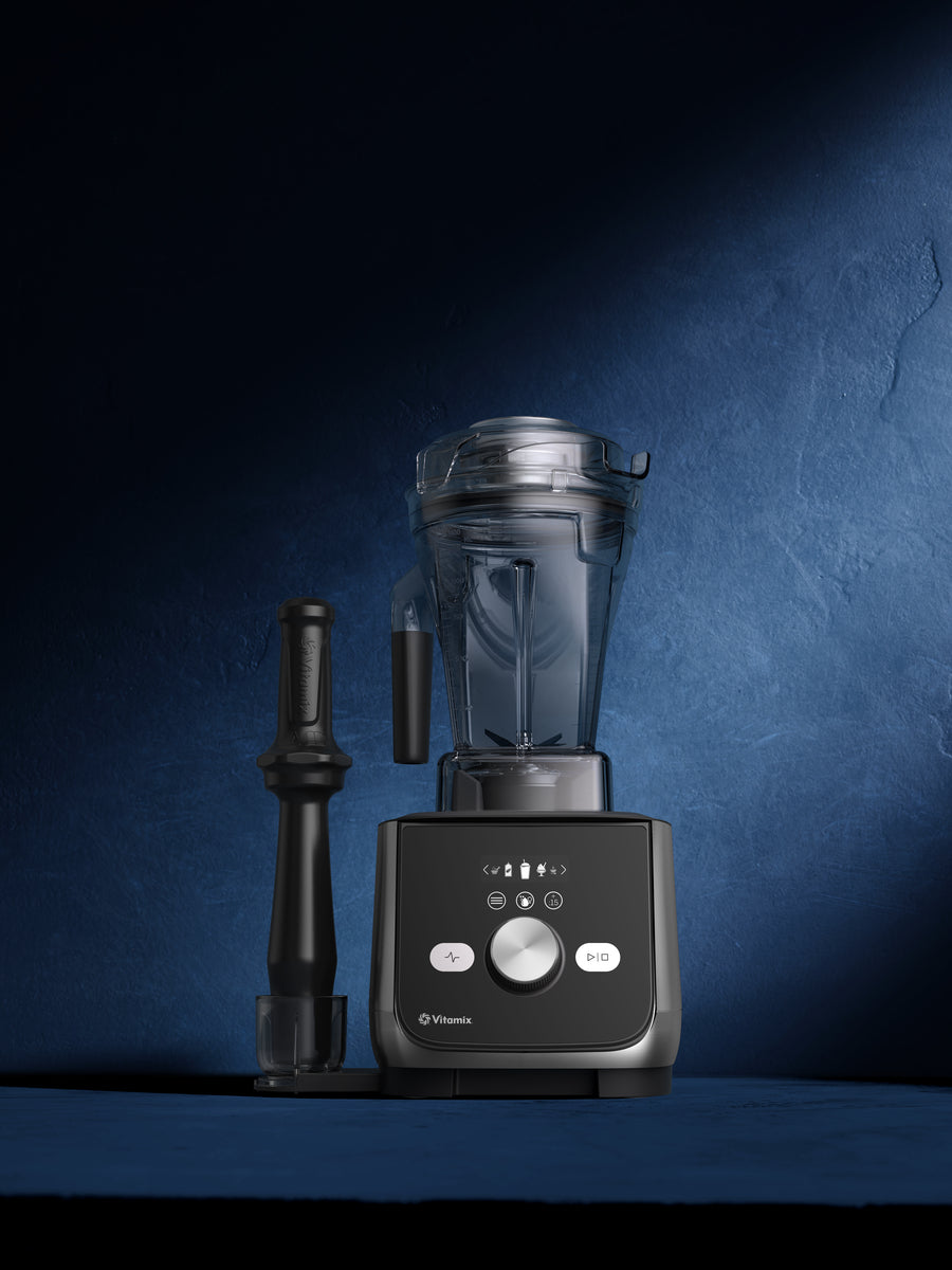 A Vitamix Ascent X5 blender with a clear container, black and gray base, and a black tamper, set against a dark blue textured background.
