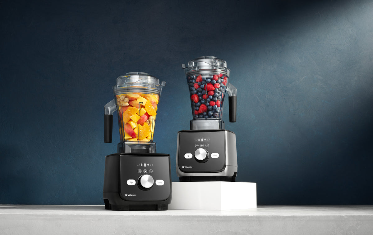 Two countertop Ascent X Vitamix blenders with clear containers are shown on a light surface against a dark blue studio background. The left blender contains orange fruit pieces, and the right blender contains red and blue berries.