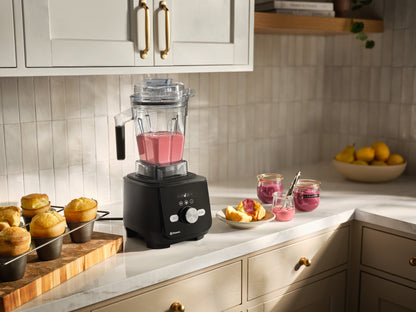 Ascent X blender filled with pink blended fruit sits on a kitchen counter, surrounded by muffins, small jars of pink jam, and a bowl of lemons. The kitchen has white cabinets, gold handles, and a light tile backsplash.