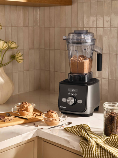 Ascent X countertop blender with a clear jug and silver and black base sits on a kitchen counter. The blender is filled with a brown icecream with bowls of dessert, a striped cloth, and a jar of coffee beans nearby against a beige tiled backsplash.