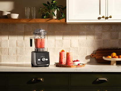 Ascent X5 countertop blender filled with pink smoothie stands in a styled kitchen setting next to two tall glasses of the same drink, with cabinets, tile backsplash, and fruit visible in the background.