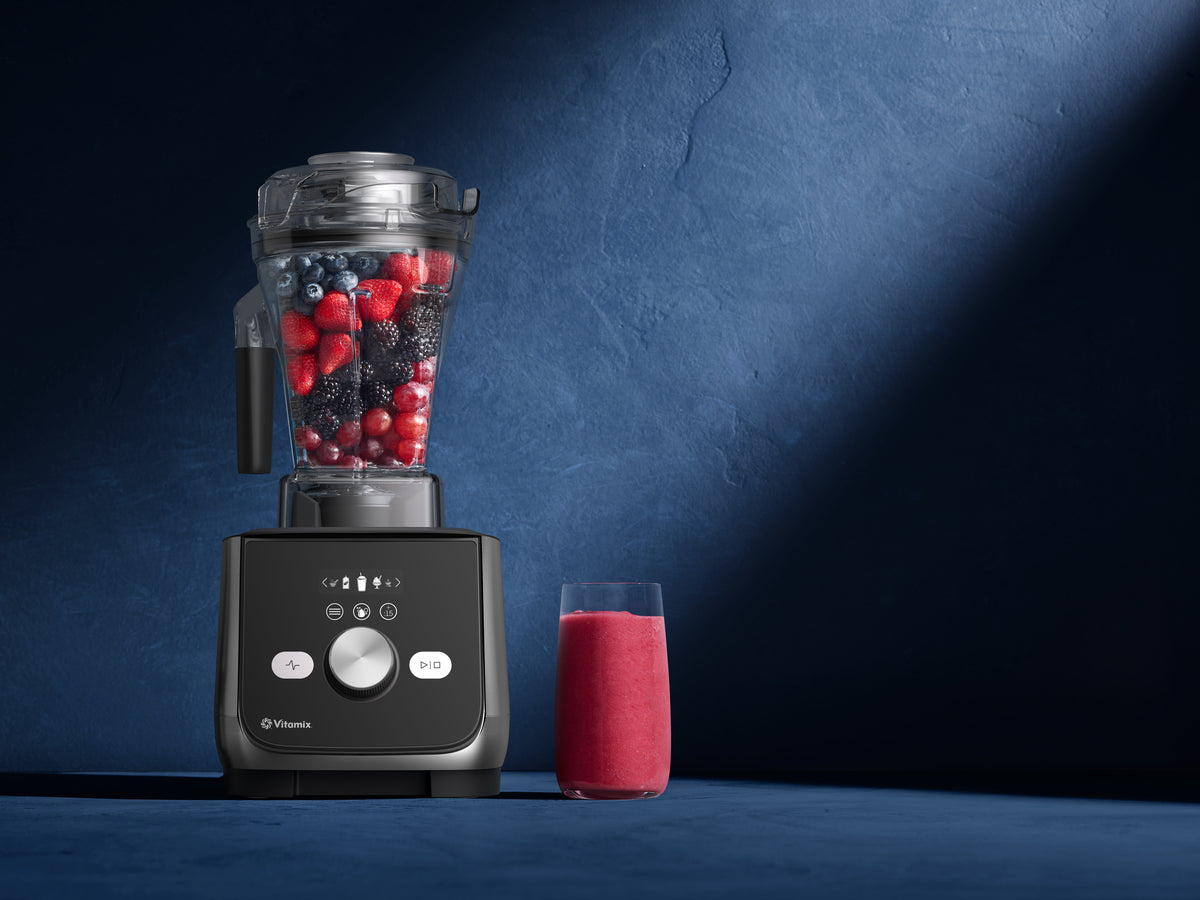 A Vitamix Ascent X blender filled with mixed berries sits on a dark blue surface next to a glass of pink smoothie against a textured blue background.