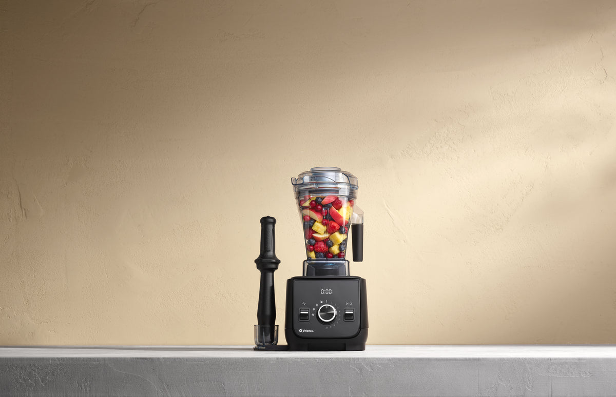 A black Vitamix Ascent X blender base with a clear container filled with chopped red and yellow fruit, standing on a light grey surface against a warm beige textured wall. A black tamper is beside the blender in its holder.
