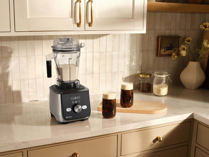 Ascent X countertop blender with a clear container and dark base sits on a light stone kitchen counter, with two frothy drinks, jars, and cream cabinets in the background.
