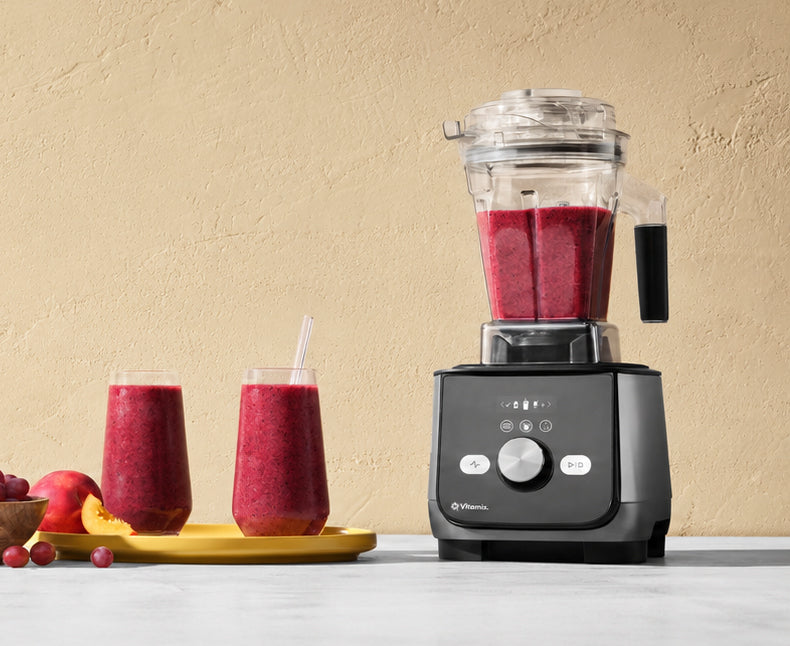 A Vitamix Ascent X5 blender with a clear container filled with a red smoothie stands on a countertop against a beige textured wall. Two glasses of the red smoothie with straws sit on a yellow tray to the left, with fruit pieces nearby.