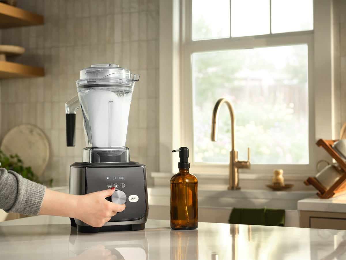 Ascent X5 blender with a clear container with bubbles inside and dark base sits on a kitchen counter, with a hand turning the dial. An amber glass bottle is beside it, and a kitchen sink and window are in the background.